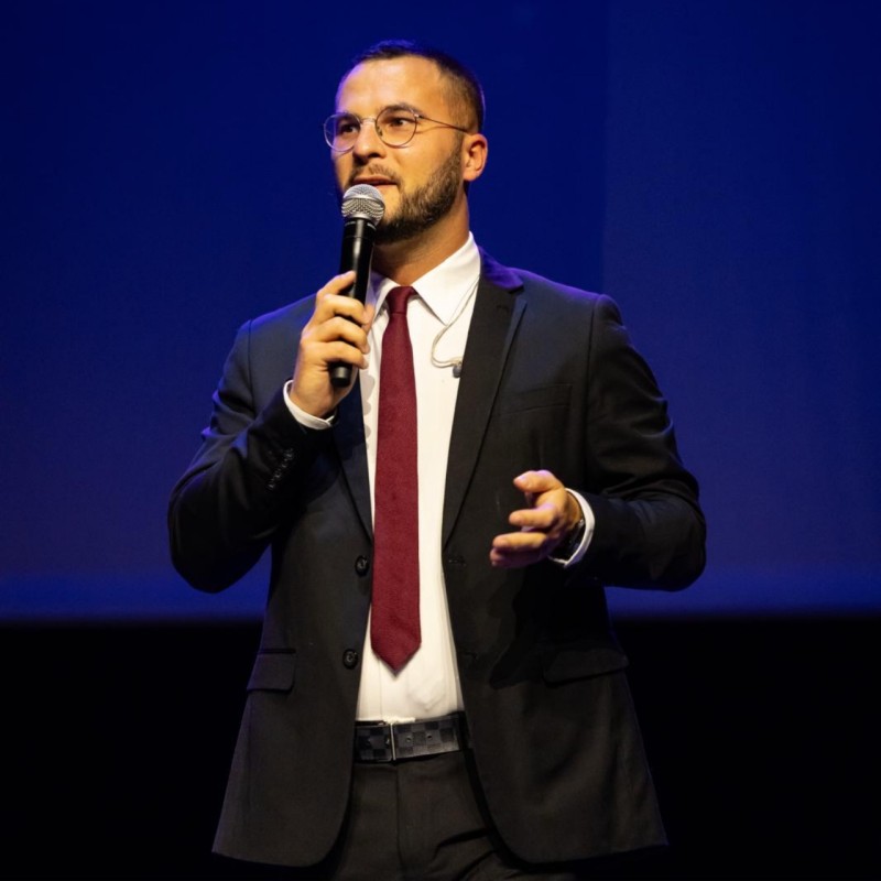 Homme en costume parle sur scène avec micro.